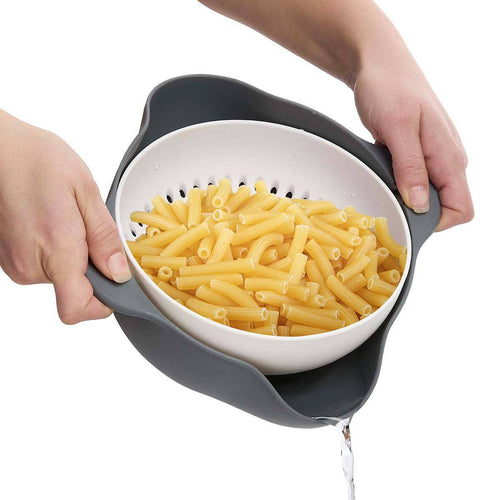 Person holding a white colander with gray handles filled with cooked pasta on a white background