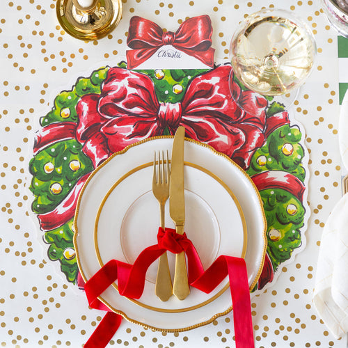 Christmas-themed place setting with a decorative plate, cutlery, and red ribbon on a red bow wreath placemat over a gold polka dot tablecloth.