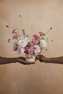 Two hands holding a bouquet of pink and white flowers in a mug labeled 'Mom' against a beige background.