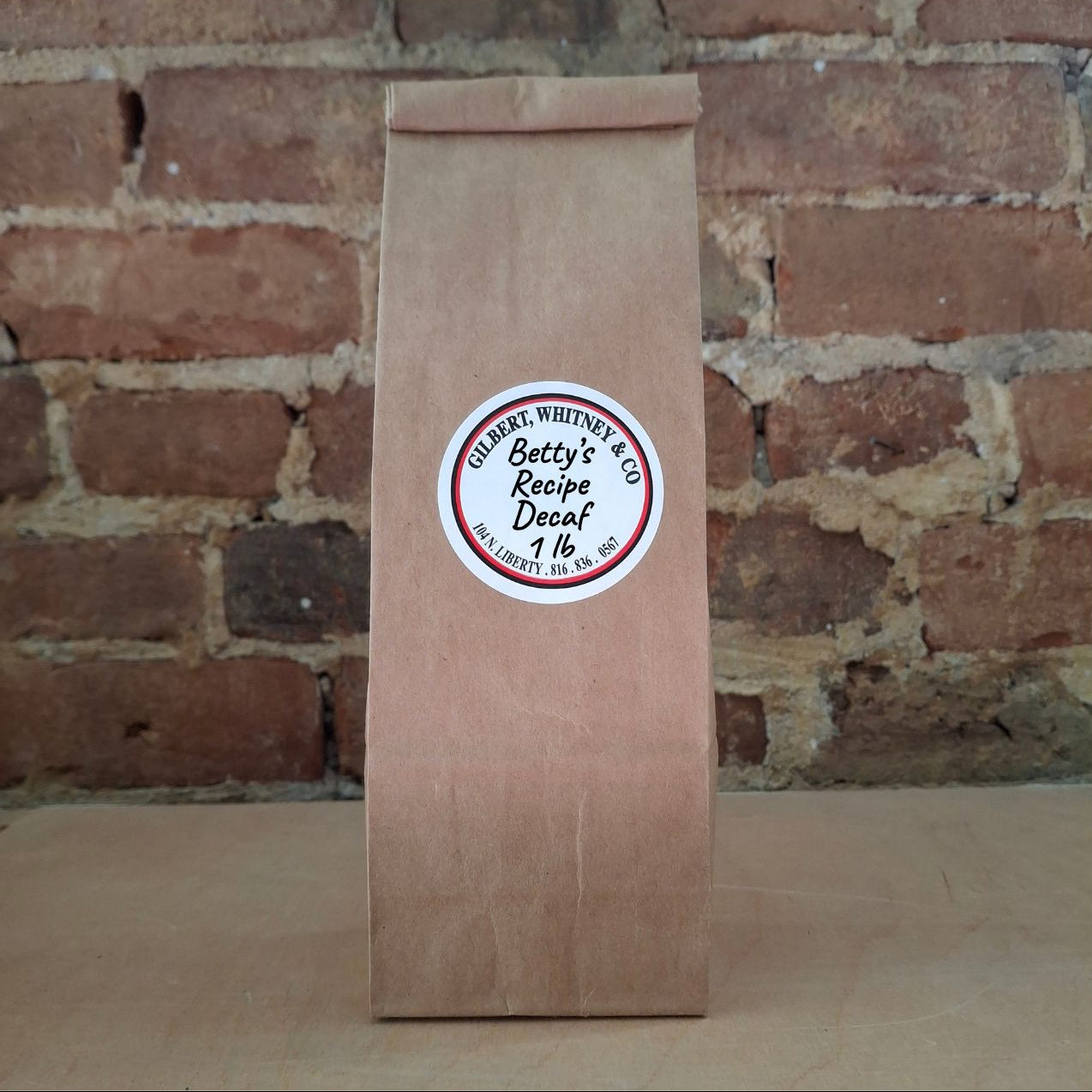 A brown kraft paper bag of coffee standing upright against a rustic brick wall and featuring a round white label reading 'Betty's Recipe'