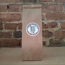 A brown kraft paper bag of coffee standing upright against a rustic brick wall and featuring a round white label reading 'Betty's Recipe'