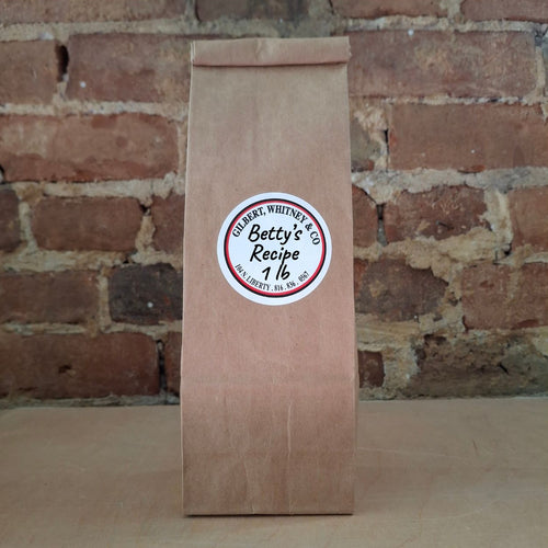 A brown kraft paper bag of coffee standing upright against a rustic brick wall and featuring a round white label reading 'Betty's Recipe'