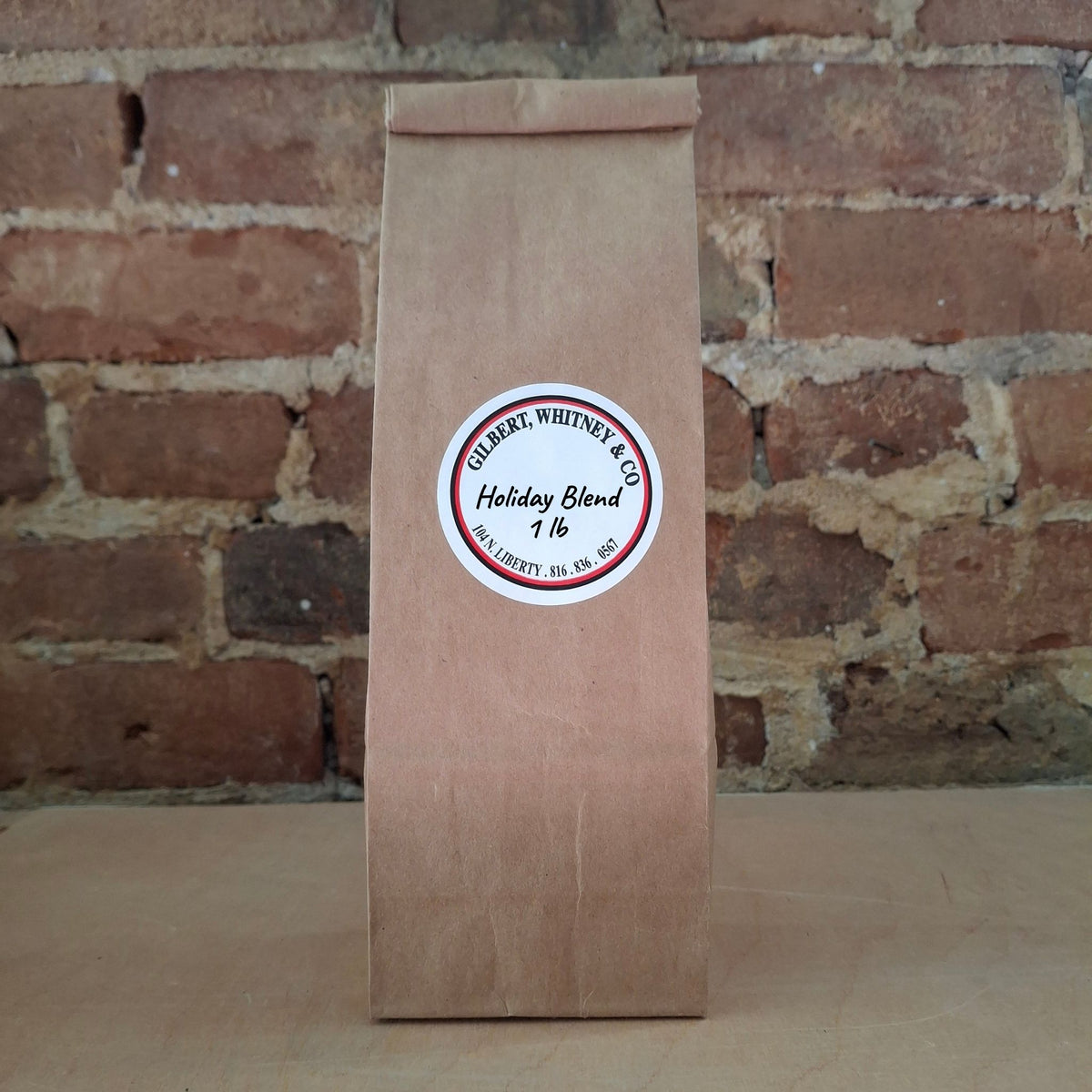A brown kraft paper bag of coffee standing upright against a rustic brick wall and featuring a round white label reading 'Holiday Blend'