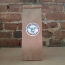 A brown kraft paper bag of coffee standing upright against a rustic brick wall and featuring a round white label reading 'Kansas City Blend Decaf'
