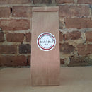 A brown kraft paper bag of coffee standing upright against a rustic brick wall and featuring a round white label reading 'Ophelia's Blend'