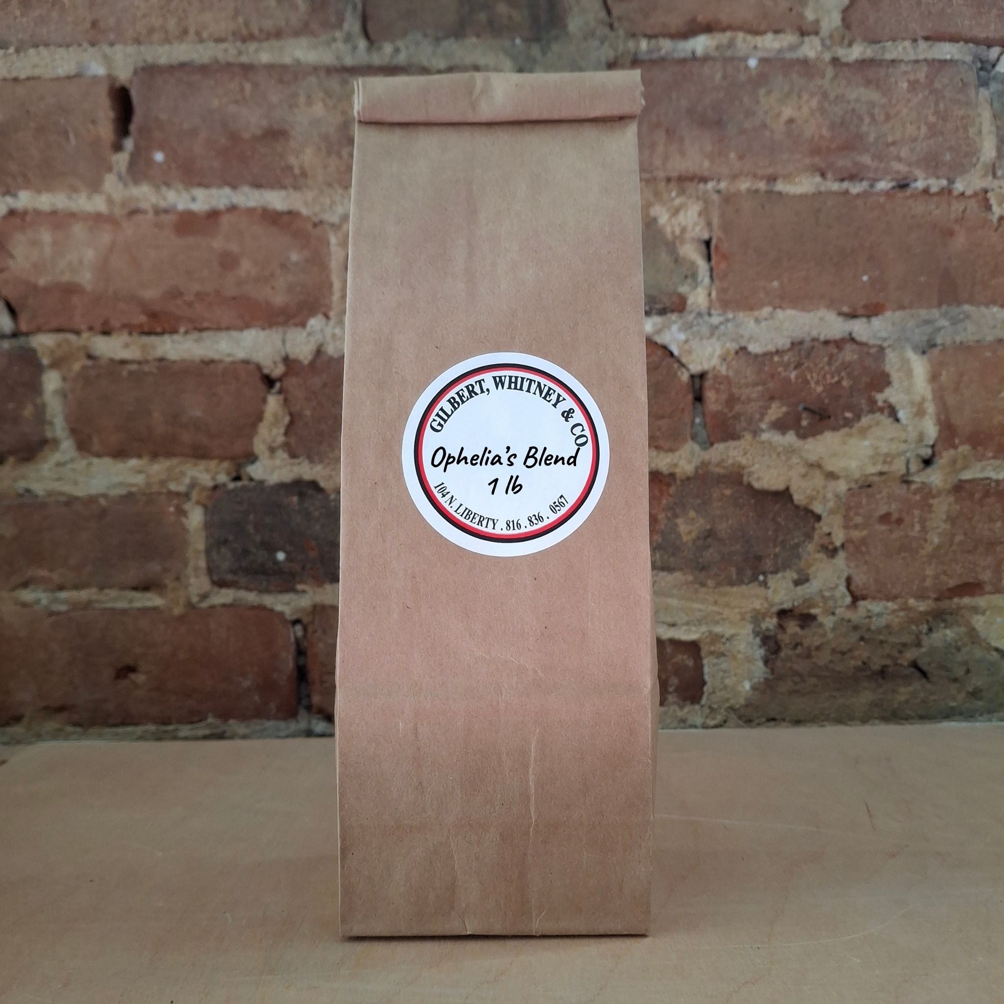 A brown kraft paper bag of coffee standing upright against a rustic brick wall and featuring a round white label reading 'Ophelia's Blend'