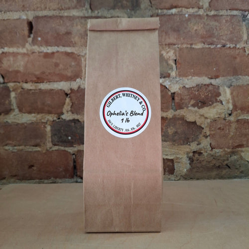 A brown kraft paper bag of coffee standing upright against a rustic brick wall and featuring a round white label reading 'Ophelia's Blend'