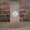 A 1 lb brown kraft paper bag of French Caramel Cream coffee, featuring a round white label with red and black text, set against a rustic red-brown brick wall background.