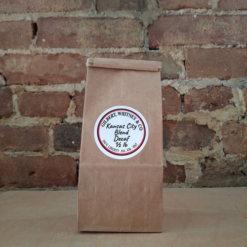 A brown kraft paper bag of coffee standing upright against a rustic brick wall and featuring a round white label reading 'Kansas City Blend Decaf'