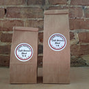 Two brown kraft paper bags of coffee standing upright against a rustic brick wall. Both bags have round white labels reading ‘Cafe Verona blend'