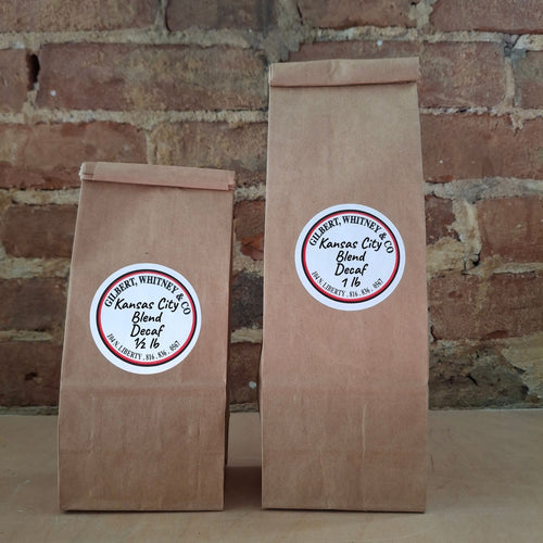 Two brown kraft paper bags of coffee standing upright against a rustic brick wall. Both bags have round white labels reading ‘Kansas City Blend Decaf'