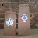Two brown kraft paper bags of coffee standing upright against a rustic brick wall. Both bags have round white labels reading ‘KC Kingdom Blend'