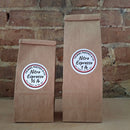 Two brown kraft paper bags of coffee standing upright against a rustic brick wall. Both bags have round white labels reading 'Nitro Espresso'