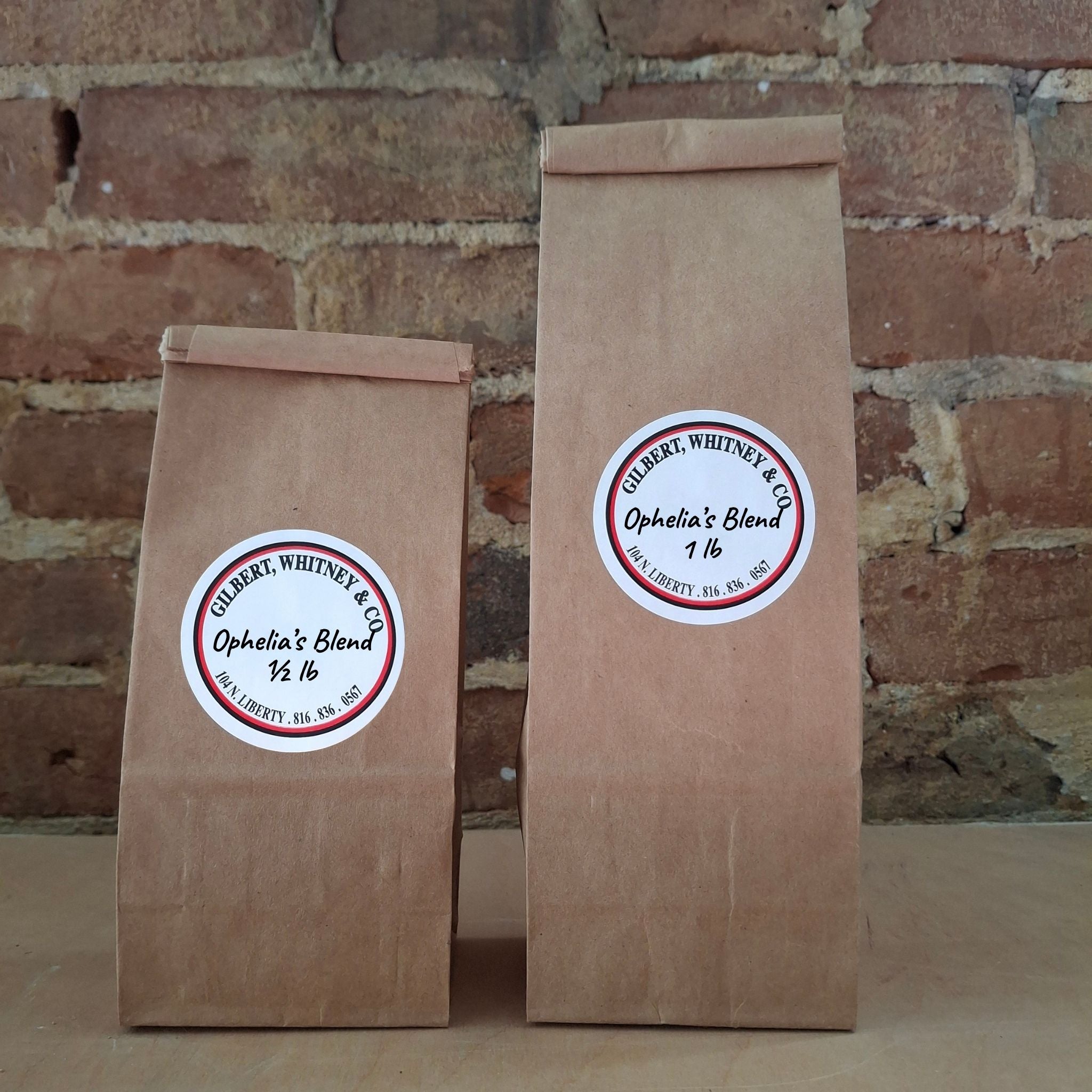 Two brown kraft paper bags of coffee standing upright against a rustic brick wall. Both bags have round white labels reading ‘Ophelia's Blend'