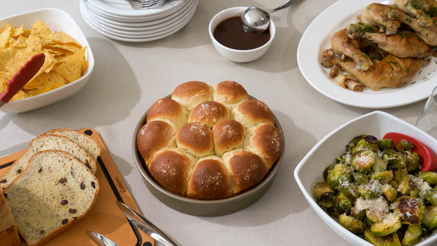 A table full of plates and bowls with food with a baking pan in the center full of bread rolls.