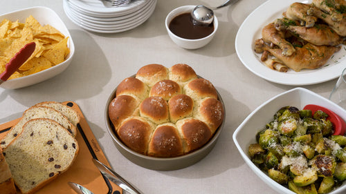 A table full of plates and bowls with food with a baking pan in the center full of bread rolls.