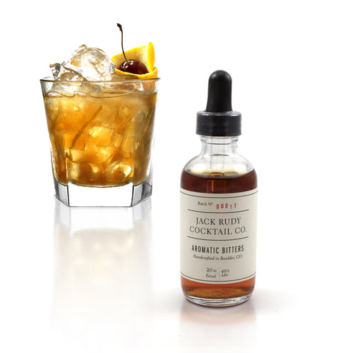 A bottle of Jack Rudy Aromatic Bitters with a dropper cap, next to a cocktail on a white background.