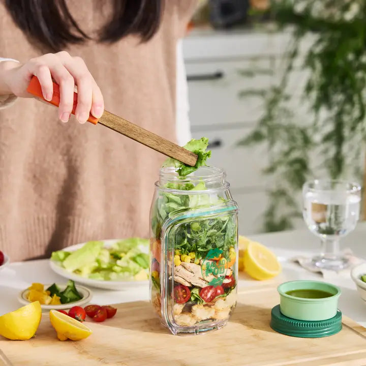 Person preparing a salad in a mason jar with a wooden spoon, surrounded by ingredients on a kitchen counter.