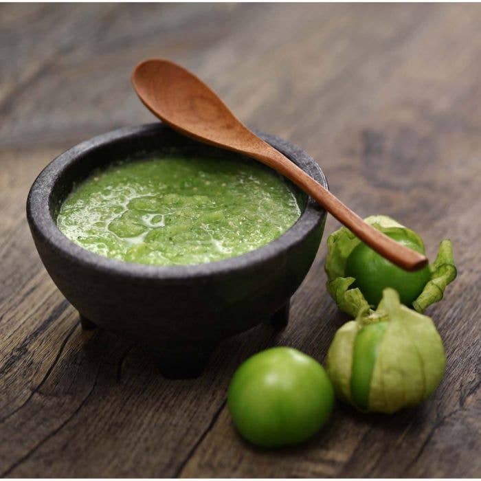 Green salsa in a black bowl with a wooden spoon on a wooden surface, accompanied by tomatillos.