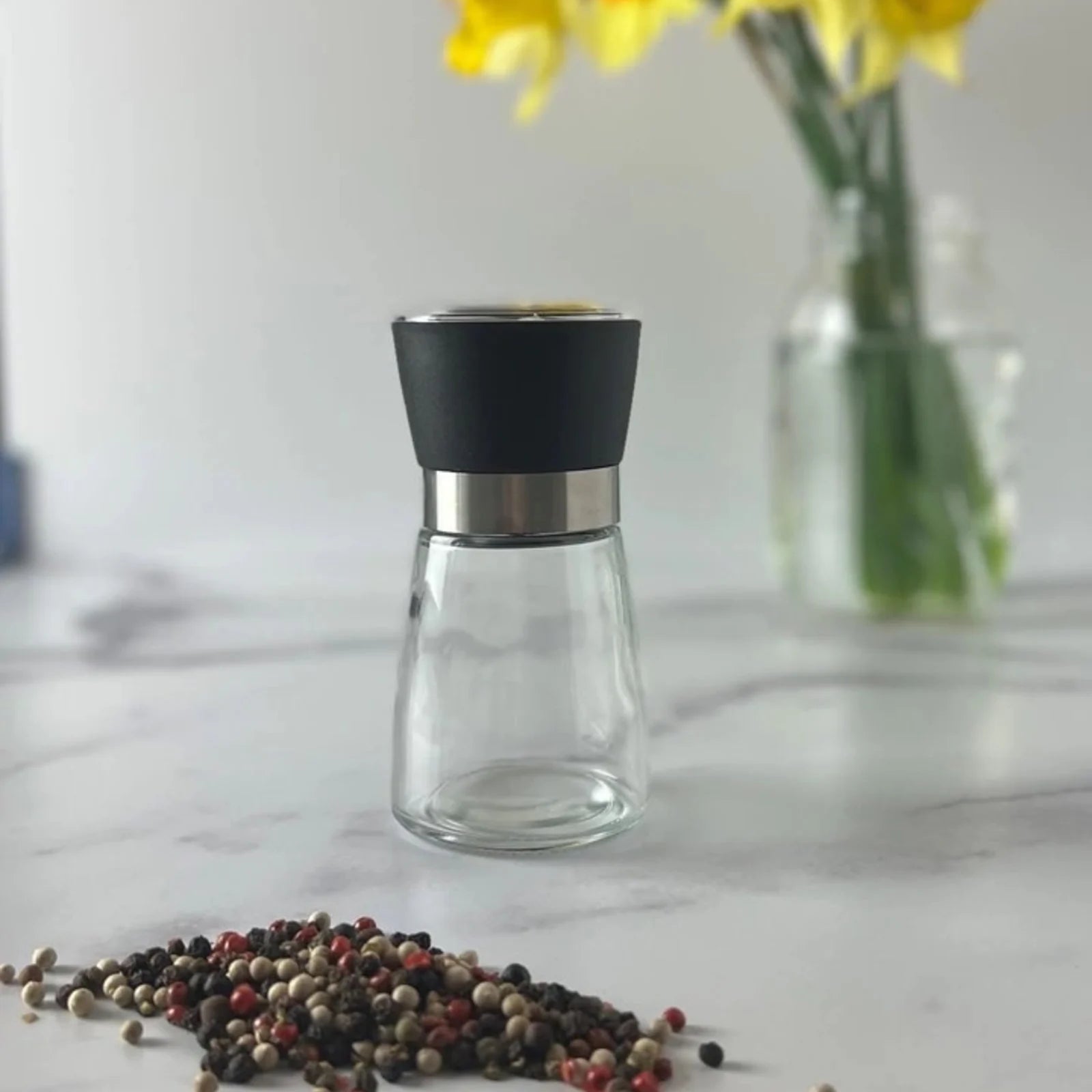 A multi purpose grinder for spices on a marble surface with a vase of daffodils behind it and a pile of peppercorns in front of it.