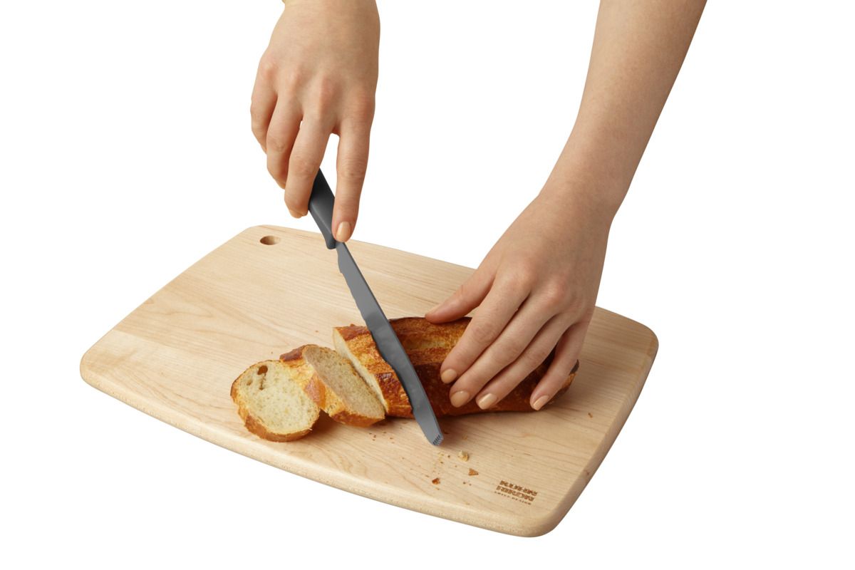 A light natural wood cutting board with rounded corners, shown on a white background with a person's hands slicing a loaf of bread with a dark-handled knife.
