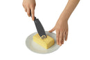 A black serrated kitchen knife with a spiral-cutting tip pressed into a rectangular block of yellow butter on a white plate, with two hands visible demonstrating use against a white background.