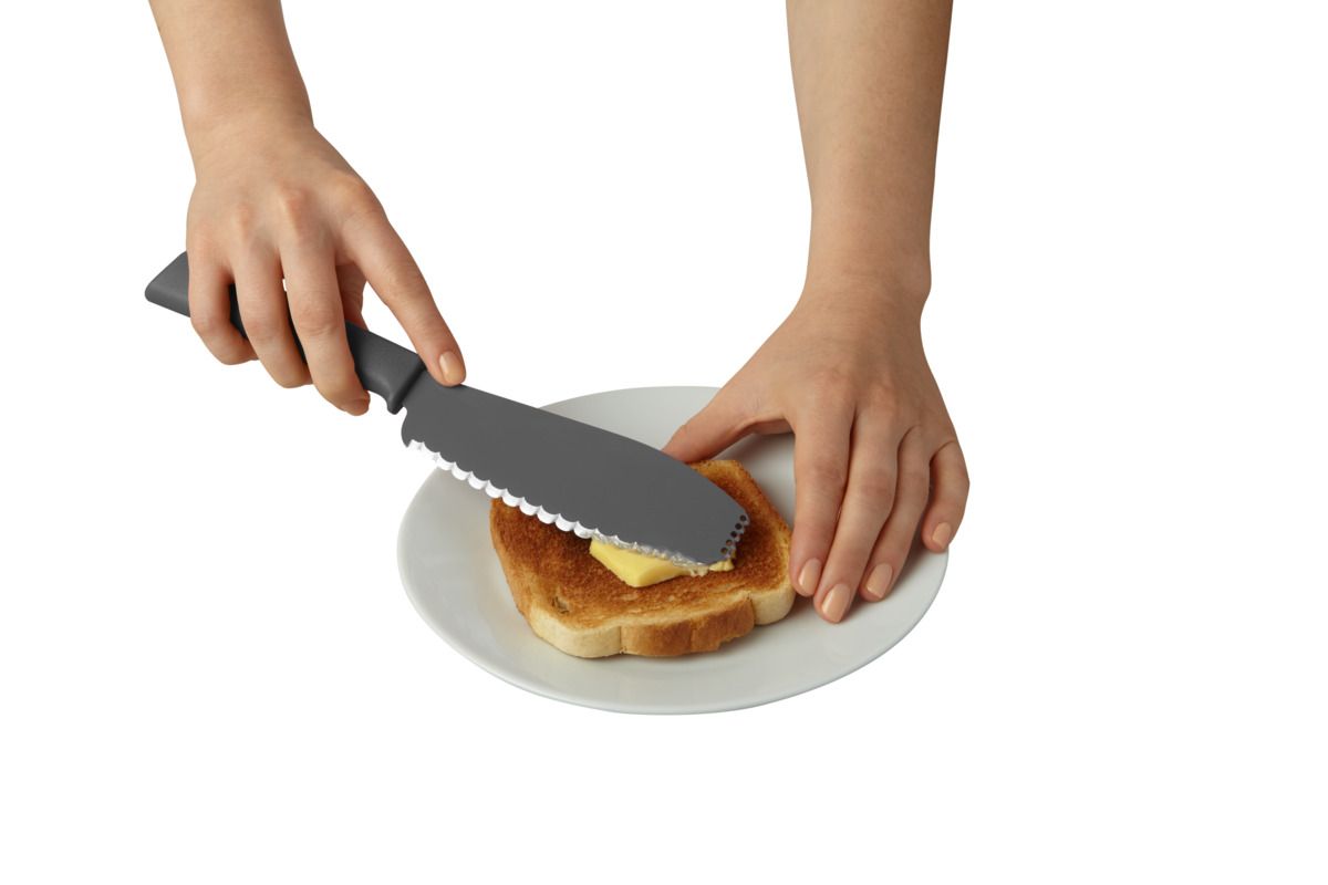 A person's hands use a large gray serrated kitchen knife to cut a slice of toast with butter on a white plate, isolated on a white background.