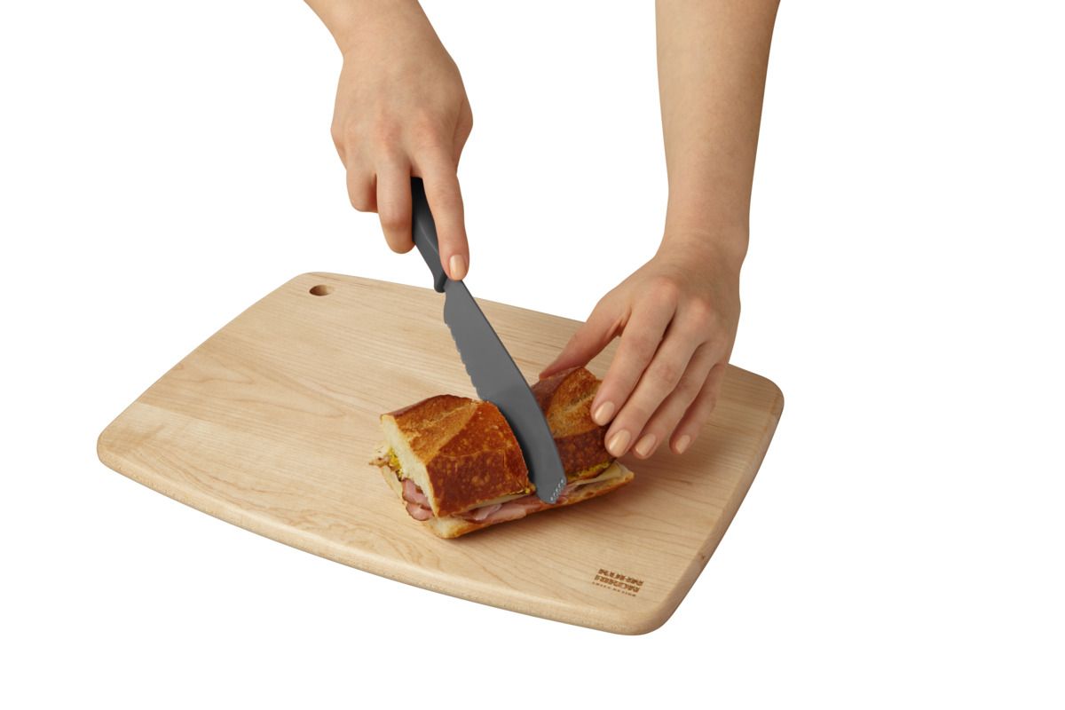 A wooden cutting board with rounded corners on a white background, featuring a person's hands slicing a sandwich with a black-handled knife.
