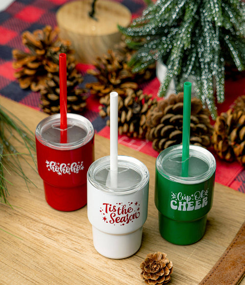 Three Christmas-themed tumblers with straws on a wooden surface with decorative elements.