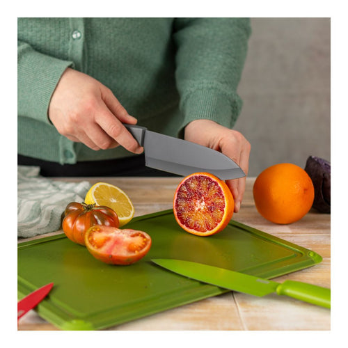 A person in a green sweater holds a dark gray kitchen knife over a green cutting board, slicing a cut blood orange. Other fruits and vegetables, including a whole orange, a halved tomato, a lemon half, and a red knife, are visible on the board and around it.