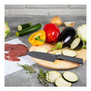 A black-handled kitchen knife with a silver blade rests on a round wooden cutting board, surrounded by fresh vegetables including eggplant, zucchini, bell peppers, cabbage, and sliced zucchini. A white cutting board with raw red meat is visible on the left, set on a gray countertop with a light gray brick wall background.
