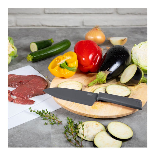 A black-handled kitchen knife with a silver blade rests on a round wooden cutting board, surrounded by fresh vegetables including eggplant, zucchini, bell peppers, cabbage, and sliced zucchini. A white cutting board with raw red meat is visible on the left, set on a gray countertop with a light gray brick wall background.