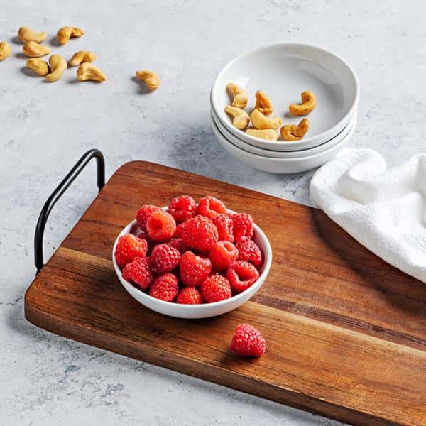 A small bowl filled with raspbrries on a wooden tray and a stack of bowls with a few cashews, all on a white surface and more cashews scattered across it.