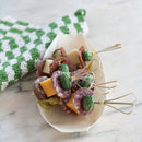 Platter of skewered appetizers on a elongated plate with a green and white checked fabric on a marble surface.