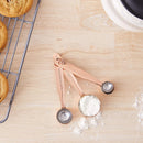 A set of copper finish measuring spoons arranged on a light wood surface, with one cup filled with white powder. Partially visible cookies on a cooling rack and a bowl appear at the edges as styled kitchen props.