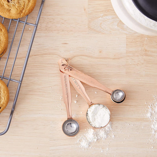 A set of copper finish measuring spoons arranged on a light wood surface, with one cup filled with white powder. Partially visible cookies on a cooling rack and a bowl appear at the edges as styled kitchen props.