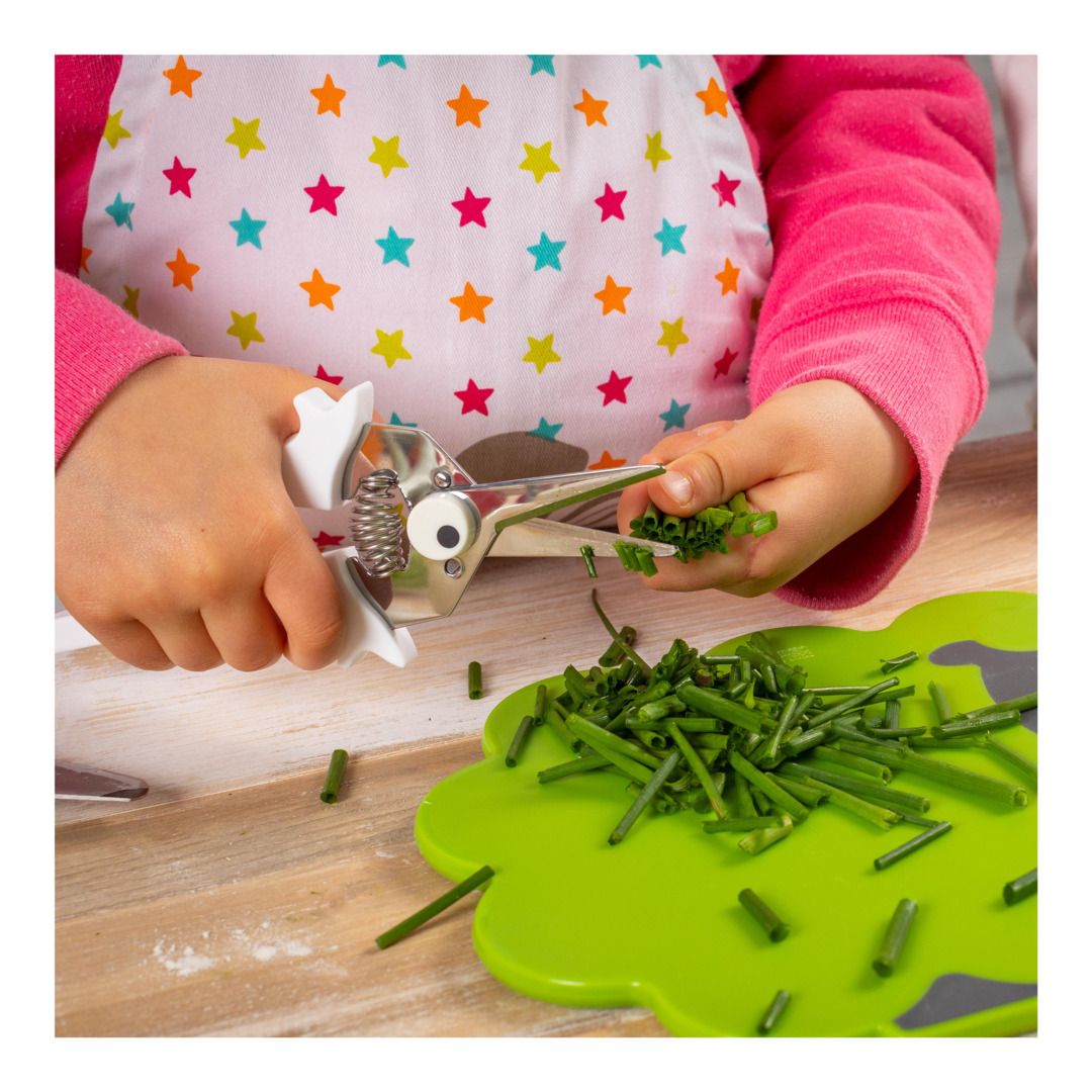 A child's hands using a metal and white vegetable chopper tool to cut green herbs over a bright green cutting board on a wooden table. The child is wearing a pink long-sleeve shirt and a white apron with colorful star patterns.
