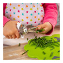A child's hands using a metal and white vegetable chopper tool to cut green herbs over a bright green cutting board on a wooden table. The child is wearing a pink long-sleeve shirt and a white apron with colorful star patterns.