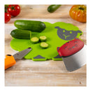 A bright green sheep-shaped cutting board with a gray sheep face, shown on a wooden surface with cucumbers and sliced pieces. A red-handled knife and an orange-handled peeler are placed around it, with tomatoes and a pumpkin blurred in the background.