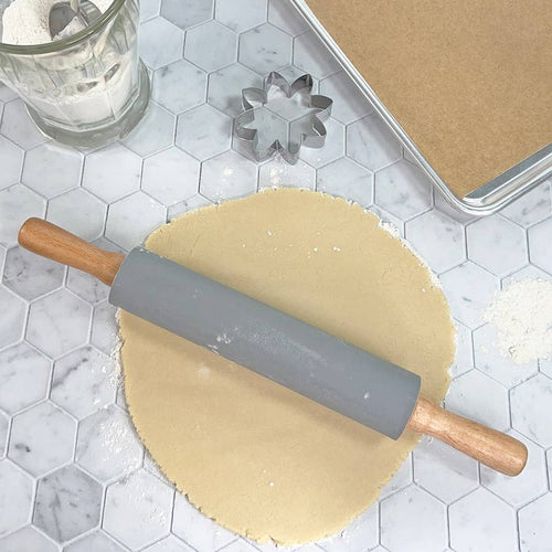 A rolling pin with a gray barrel resting atop a circle of dough near a cookie cutter and baking pan all on a gray surface of hexagonal tiles.