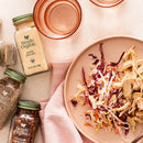 A styled flat-lay featuring Simply Organic spices, including garlic powder, ground pepper, and dried herbs, alongside a plate of shredded chicken and coleslaw on a pink cloth over a light beige surface.