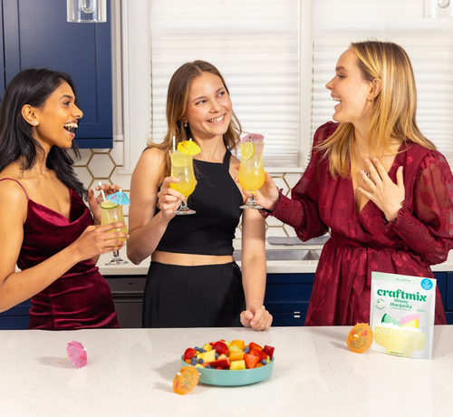 Three women in a kitchen enjoying drinks with a bowl of fruit and a box of craftmix on the counter.