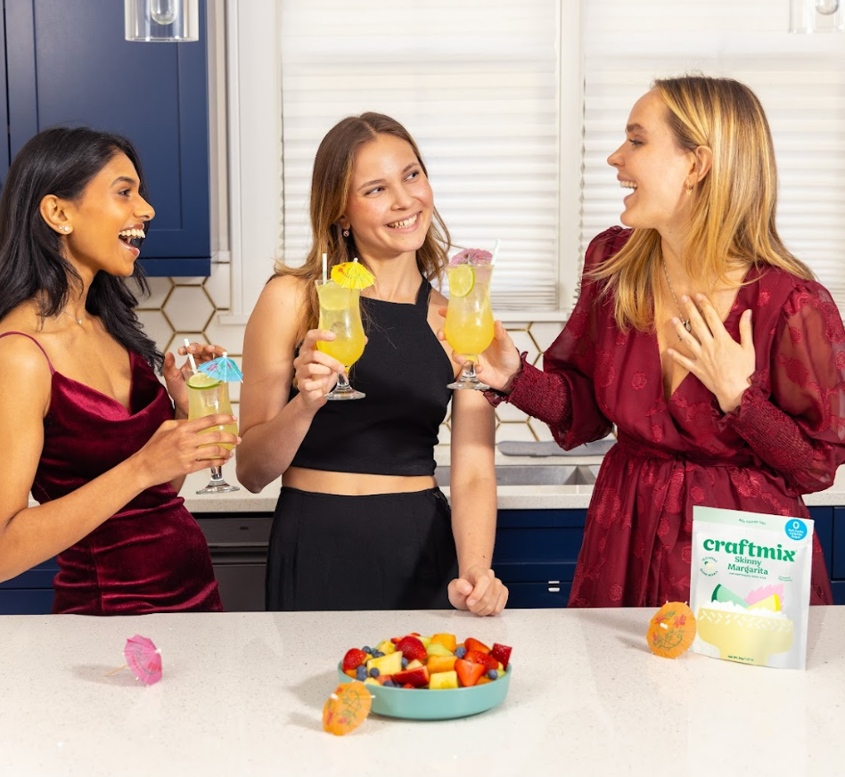 Three women in a kitchen enjoying drinks with a bowl of fruit and a box of craftmix on the counter.