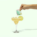 Hand pouring a packet into a margarita with a lime wedge on a white background