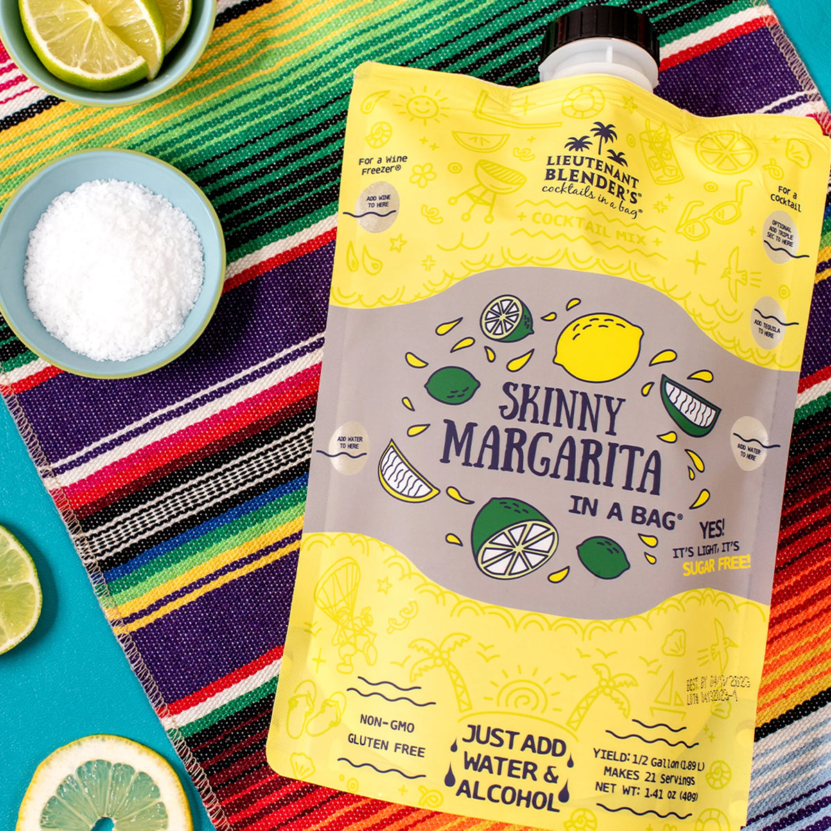 Yellow 'Skinny Margarita in a Bag' package on a colorful striped towel with lime slices and salt.