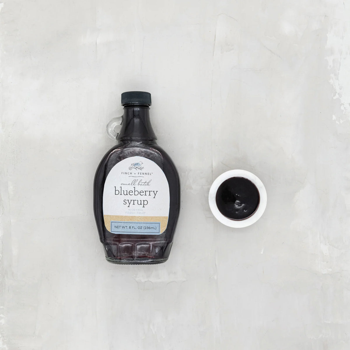 Bottle of blueberry syrup with a small bowl of syrup on a textured white background