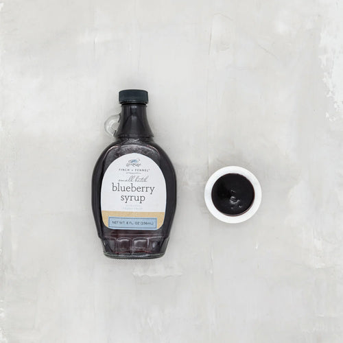 Bottle of blueberry syrup with a small bowl of syrup on a textured white background