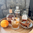 A styled cocktail scene on a round woven tray with a glass of amber drink, an orange, cinnamon sticks, a bottle of Vermont maple syrup, a bottle of dark spirit, and a metal shaker against a gray stone wall.