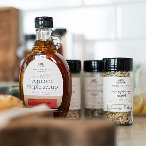 A bottle of Finch + Fennel Vermont maple syrup stands on a countertop, with several Finch + Fennel spice jars behind it in a softly blurred kitchen setting.