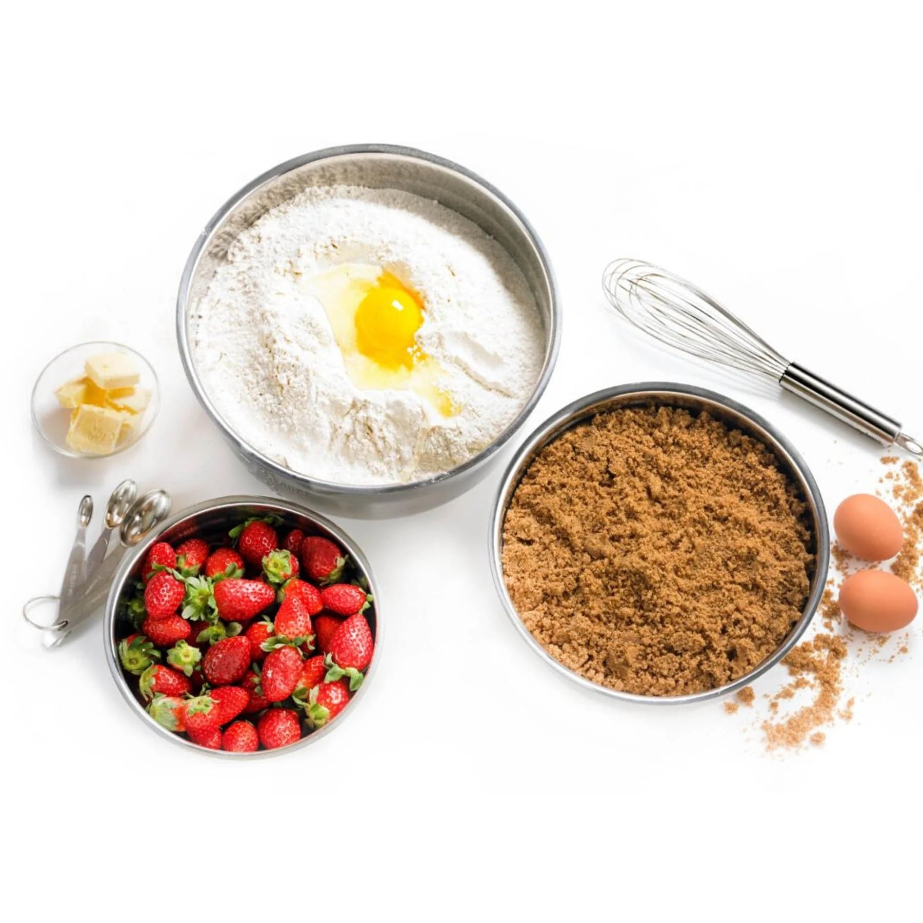 Bowls of flour, strawberries, brown sugar, and eggs on a white background with a whisk.